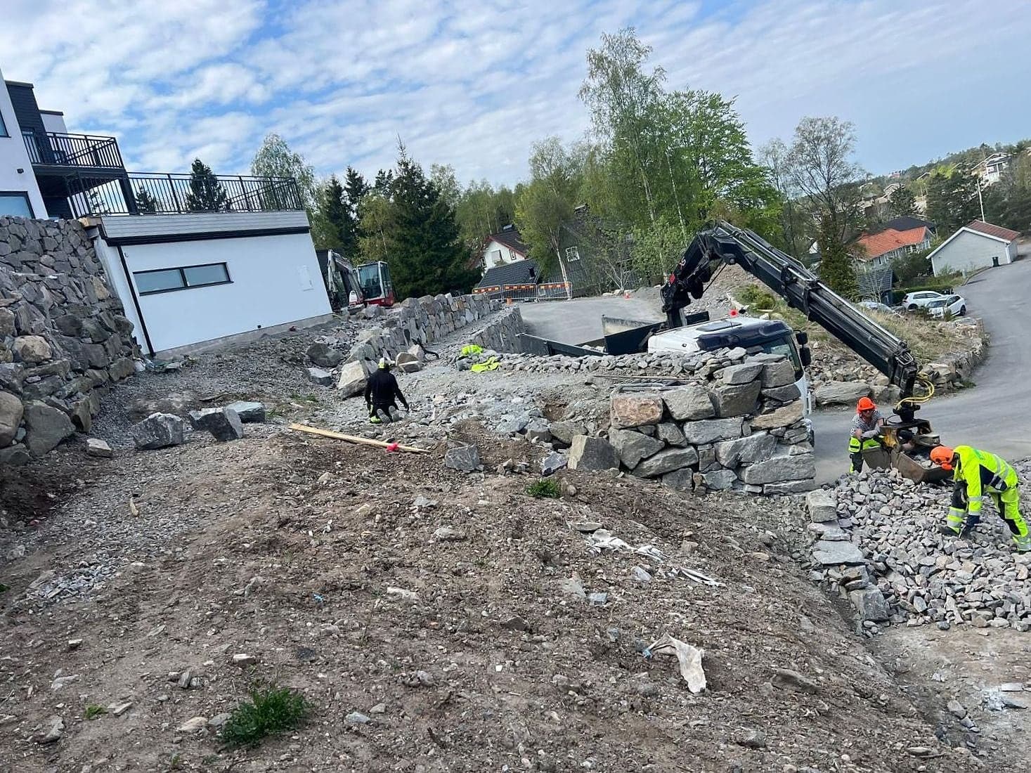 Kranbil lyfter natursten på plats medan tre arbetare i varselkläder bygger en mur.